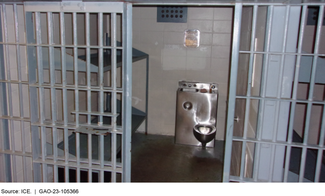 A prison cell with two empty bunks and a metal sink and toilet station. The photo is taken from outside the bars to the cell.