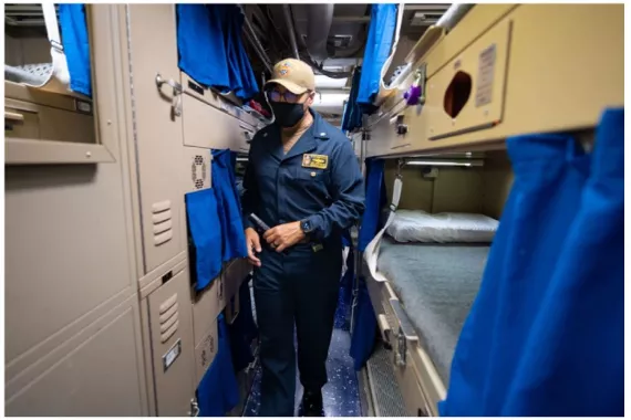 Photo showing a Navy officer walking down a hallway that has bunk beds installed on either side with little curtains.