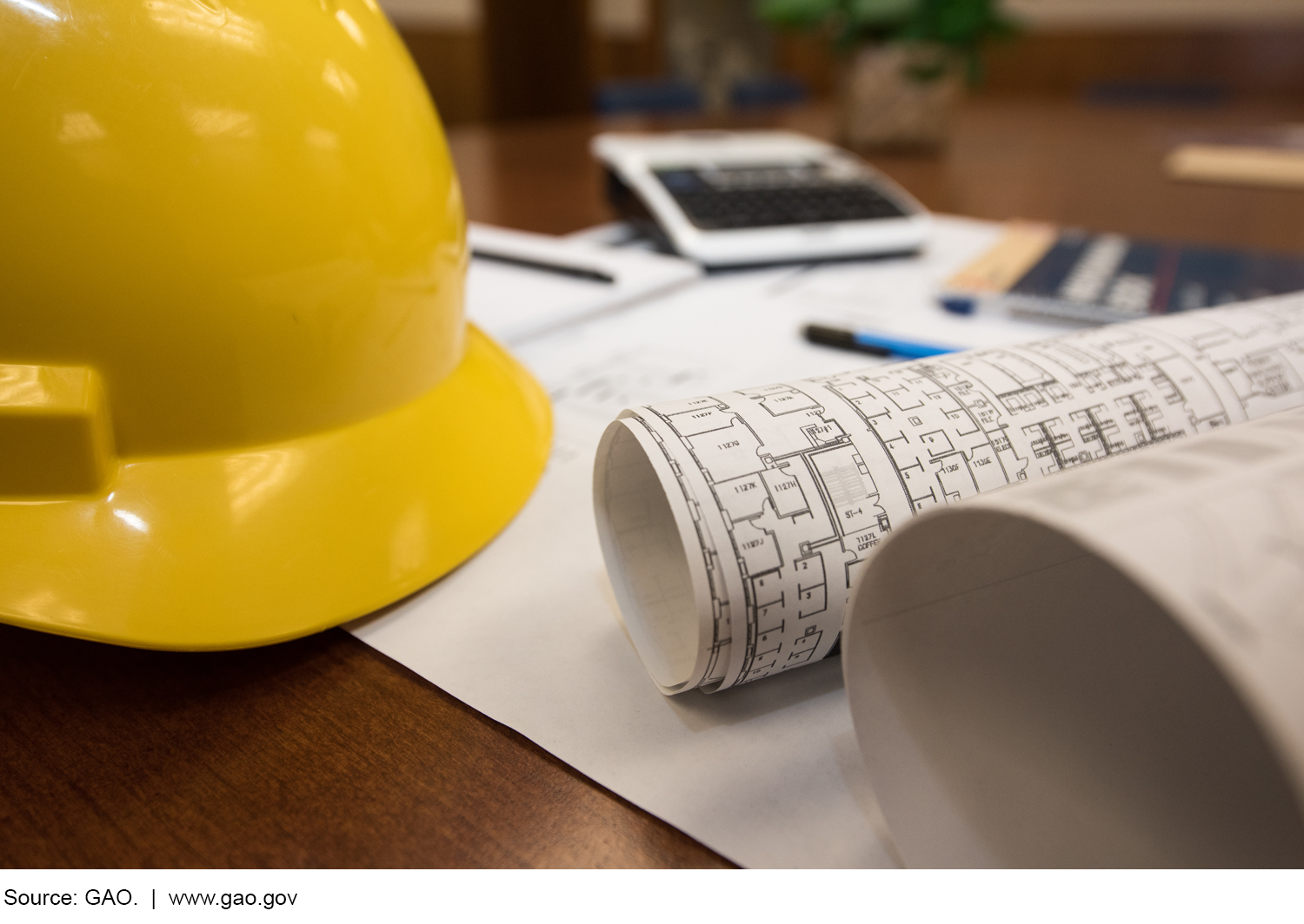 Photo of hard hat, construction plans, papers and pens on a desk.