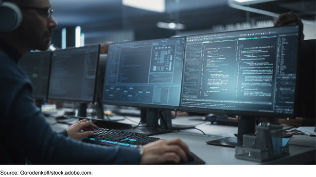 A man wearing headphones works at a computer keyboard in front of three monitors displaying computer code and other data. A man wearing headphones works at a computer keyboard in front of three monitors displaying computer code and other data.