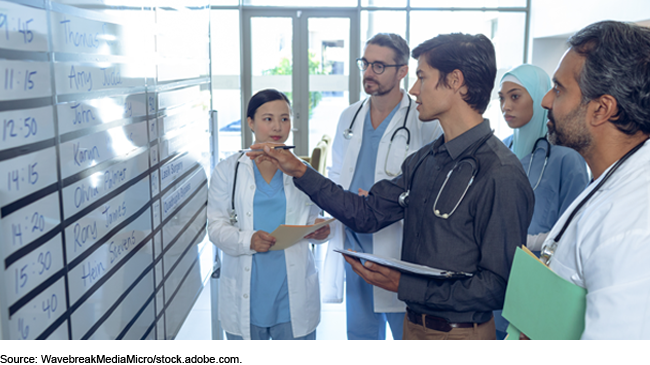 A group of people who all have stethoscopes draped around their necks look at a schedule written on a large whiteboard. Some are wearing scrubs, lab coats, or both. The person talking is wearing a dress shirt.
