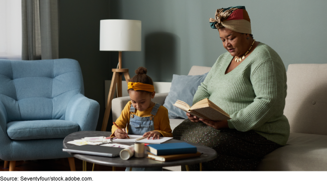 A grandmother sits reading while her granddaughter writes in a notebook near her on a coffee table.