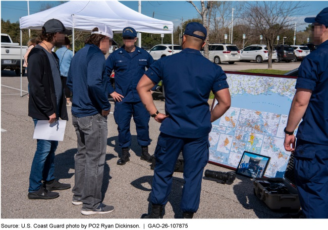 A group of people gathered on an outside parking lot on a sunny day. Coast Guard personnel in dark blue uniforms are talking to people. A large map is propped up on the ground. A group of people gathered on an outside parking lot on a sunny day. Coast Guard personnel in dark blue uniforms are talking to people. A large map is propped up on the ground.