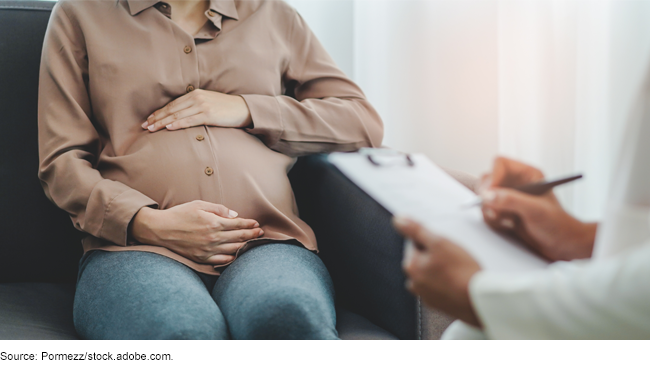 A pregnant person holding their belly while sitting across from a physician holding a clipboard and taking notes.