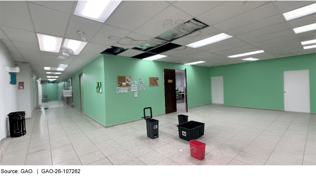 An open space with tile flooring and visible water damage on the drop ceiling above. Some ceiling tiles are missing, and the floor has a few plastic bins on it to catch water.