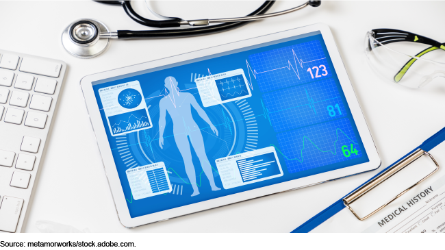 A photo that includes a stethoscope, computer keyboard, clipboard with a medical history, and a tablet computer showing patient vitals