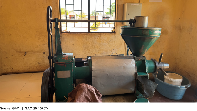 A mechanical seed oil press—a metal machine with a flywheel, a large funnel, and a valve, next to a small tub with a bucket in it. A mechanical seed oil press—a metal machine with a flywheel, a large funnel, and a valve, next to a small tub with a bucket in it.