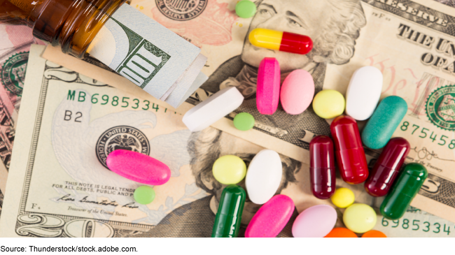 Stock photo of a pile of cash with an assortment of multicolored pills scattered on top. A rolled up $100 bill pokes out of the neck of a brown glass bottle, lying on its side on top of the other money.