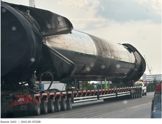 Very large metal rocket booster being towed on a long flatbed truck