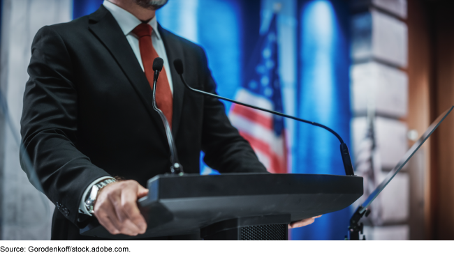 A person standing at a podium with the U.S.A. flag in the background.