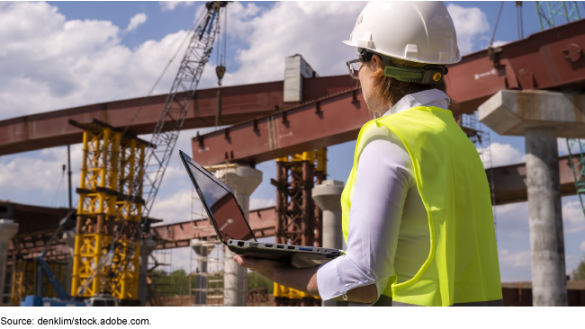Worker at a construction site wearing a yellow vest and a hard hat, while holding a laptop