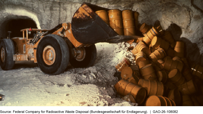 Inside an underground chamber with rock walls, a large industrial vehicle moves several yellow cylindrical drums that resemble barrels to a pit that is partially filled with other drums. Inside an underground chamber with rock walls, a large industrial vehicle moves several yellow cylindrical drums that resemble barrels to a pit that is partially filled with other drums.