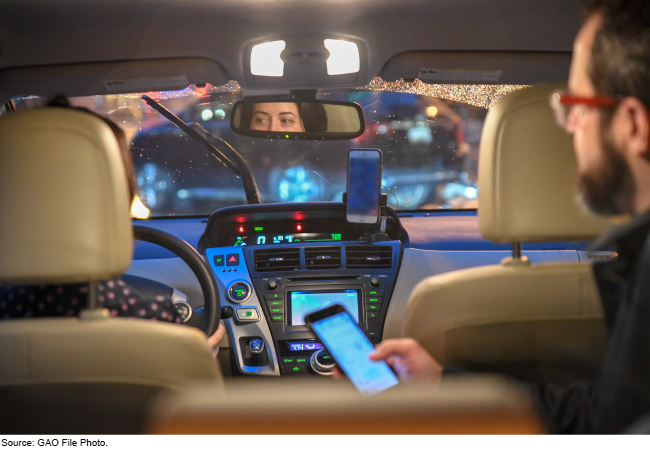 Interior of a car with a person driving and a passenger in the back seat holding a cell phone.