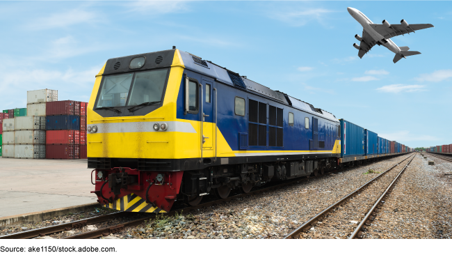 A freight train stopped alongside a platform with shipping containers stacked up on it and an airplane overhead