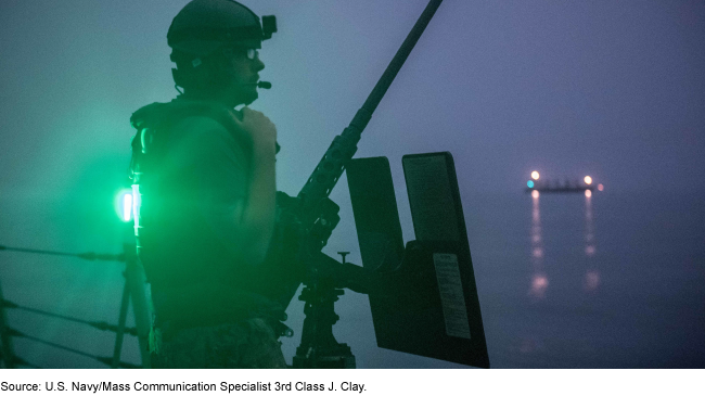 U.S. Navy crewmember standing watch at night, with a ship in the water in the distance.