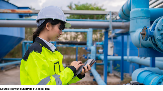 Woman wearing hard hat and safety jacket standing near wastewater system checking a tablet.