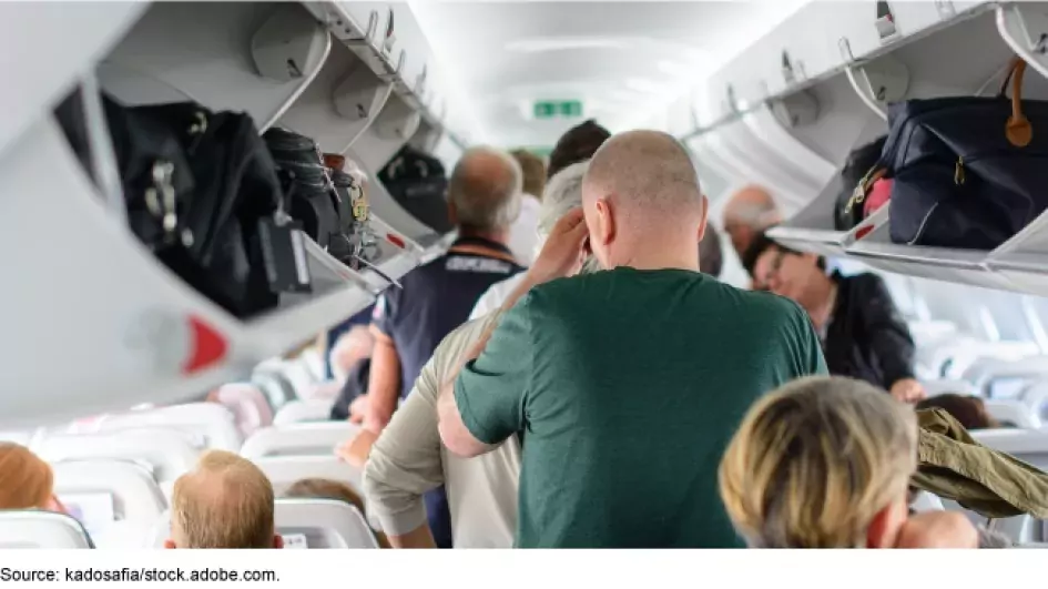 Passengers wait to exit an airplane, some sitting and some standing in the aisle. The overhead bins are open and full of luggage.