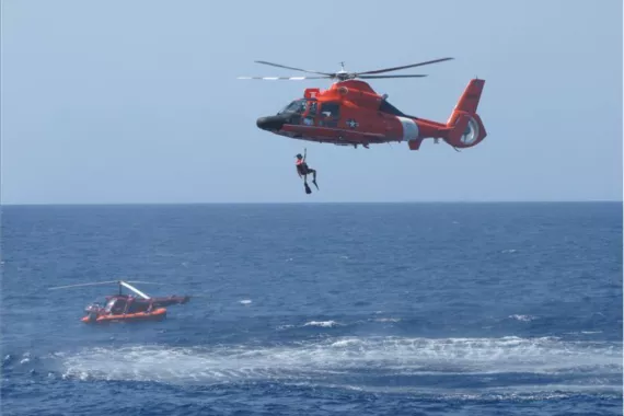 Photo showing a Coast Guard helicopter hovering over the ocean while raising a person from the water.